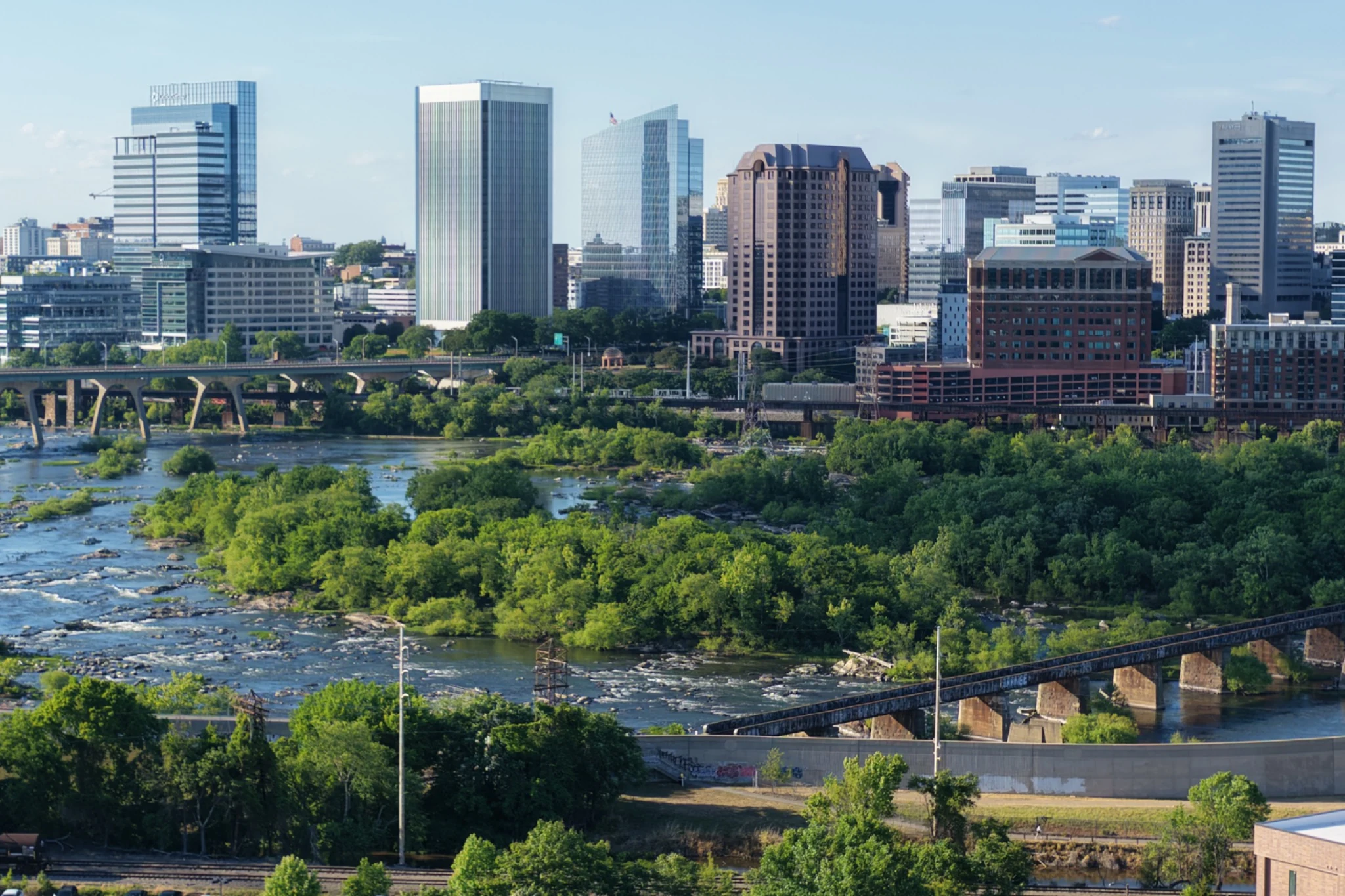 Daylight aerial of the Richmond skyline, James River, bridges, rail corridor and tree canopy.